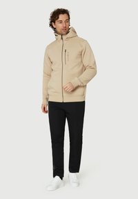 Sudadera beige con cremallera en tejido suave, con bolsillos tipo canguro y un alto cuello, combinada con pantalones negros y zapatillas blancas.