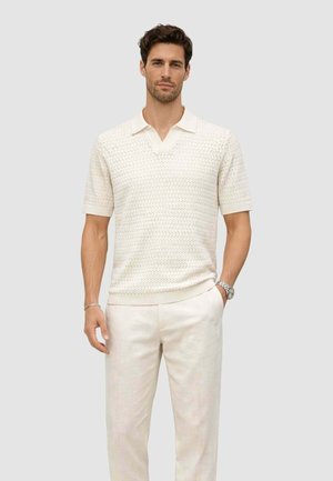 Homme portant un polo en maille à manches courtes texturé crème et un pantalon léger assorti, debout avec une main dans la poche devant un fond uni.