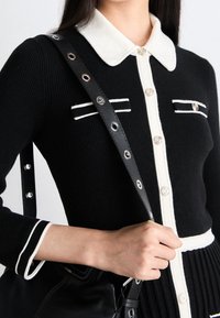 Cardigan noir à côtes avec des accents blancs, fermeture à boutons, et deux poches sur la poitrine. Sac noir avec des finitions en argent, porté à l'épaule.