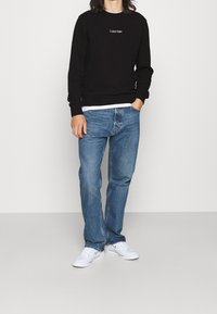 Persona che indossa un felpa nera Calvin Klein, jeans blu e sneakers bianche Nike, in piedi con una mano in tasca su uno sfondo neutro.