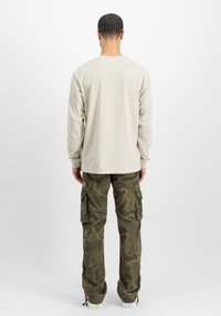 Beige shirt met lange mouwen gecombineerd met groene camouflage cargobroek met zijzakken, losse pasvorm en omgeslagen zoom, tegen een effen achtergrond.