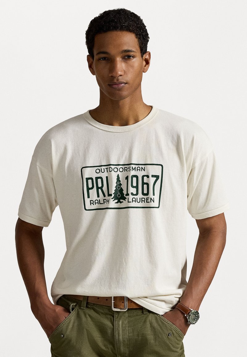 Crème katoenen t-shirt met korte mouwen, voorzien van een groene bedrukking met "OUTDOORSMAN PRL 1967" en een boomicoon. Draagt het met groene broek.