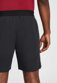 Calções atléticos pretos com cintura elástica, textura suave, comprimento até ao joelho e ajuste slim; mão repousando ao lado.