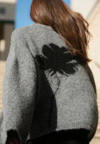 Grau gestrickter Pullover mit lockerer Passform, der ein auffälliges schwarzes Blumenmuster auf dem Rücken zeigt. Weicher Stoff und weite Ärmel erhöhen den Komfort.
