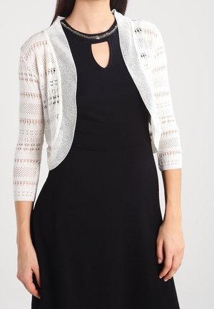 Femme portant une robe noire avec un décolleté en trou de serrure et un cardigan blanc en crochet ouvert devant avec des manches trois-quarts.