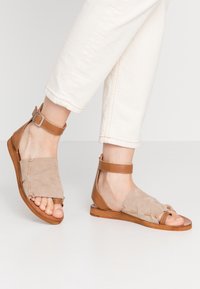 Tan suede and leather sandals with ankle strap, open toe design, flat sole, and decorative stitching details.