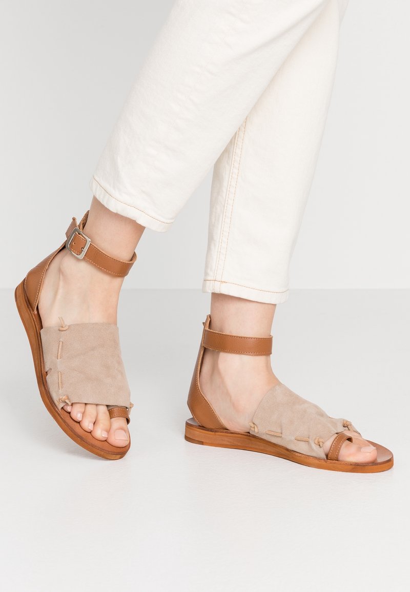 Tan suede and leather sandals with ankle strap, open toe design, flat sole, and decorative stitching details.