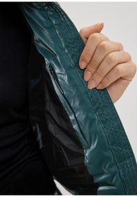 Intérieur d'une veste imperméable bleu sarcelle avec une poche zippée. Texture en plastique lisse, coutures visibles et coupe ajustée pour le confort.
