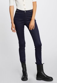 Pantalones ajustados de color navy confeccionados en tejido elástico, con bolsillos con cremallera y un diseño clásico de cinco bolsillos, combinados con botas negras hasta el tobillo.
