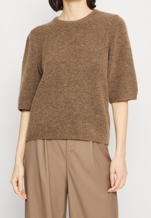 Vrouw draagt een bruin, zachte korte mouw trui en losse beige broek, staand tegen een eenvoudige lichte achtergrond.