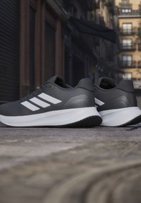 Paire de chaussures de sport gris foncé avec des rayures blanches et des semelles sur un pavement en pierre dans un environnement urbain.