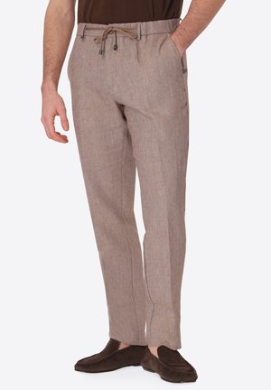 Uomo che indossa pantaloni di lino marrone chiaro con coulisse in vita e mocassini marrone scuro, mani rilassate, una in tasca.