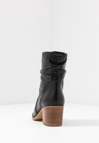 Bottine noire avec une tige en cuir texturé, des accents bouclés, et un talon en bois empilé. Design doux et froissé à l'arrière.