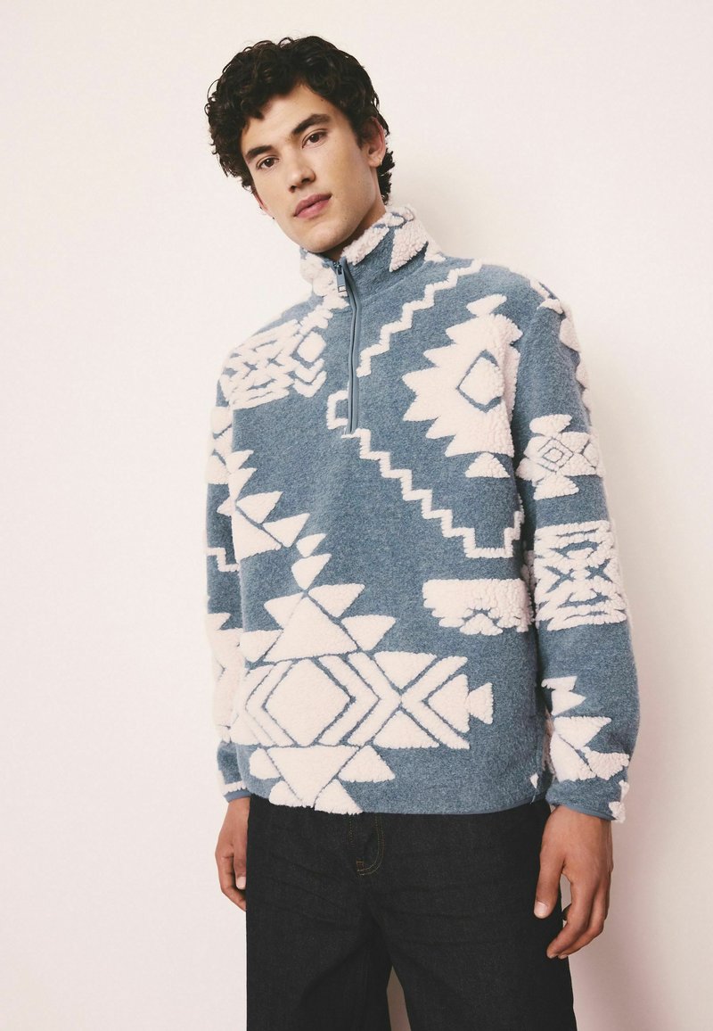 Fleece pullover in blue with white geometric patterns, zip neck, and high collar. Soft texture, long sleeves, and relaxed fit.
