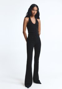 Nő ujjatlan, mély V-nyakú fekete csillogó pántos felsőben és hozzáillő, magas derekú, bő szárú nadrágban, kezét a háta mögött tartva áll.