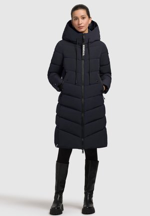 Cappotto puffo blu navy con cappuccio, design a cuciture diagonali, zip frontale, lacci e due tasche laterali. Abbinato a stivali neri fino al ginocchio.