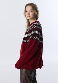Pull en tricot rouge profond avec un motif géométrique noir et blanc au niveau de l'empiècement. Texture douce, coupe oversize et poignets côtelés.