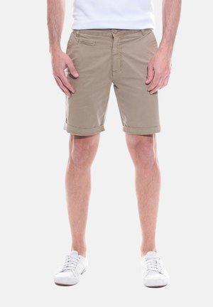 Homme portant un short beige mi-cuisse, des baskets blanches et une chemise blanche, debout devant un fond clair uni.