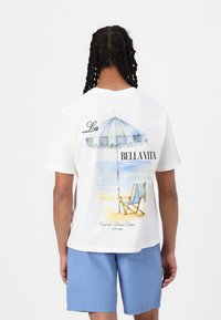 Camiseta blanca de algodón con una impresi�ón de una escena de playa que incluye una sombrilla y una silla. El texto incluye "Bella Vita" y "Edición de Verano Originales".