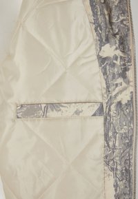 Tikkitettu untuvatakki vaaleanbeigenä, jossa harmaa maastokuvioitu vuori. Siinä on pieni ulkopuolinen tasku ja sileä, kiiltävä pinta.