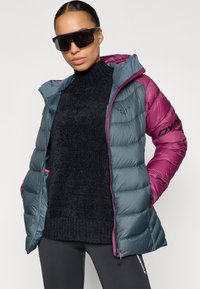 Μπουφάν με επένδυση σε τιρκουάζ και ματζέντα, με κουκούλα, δύο τσέπες και μαύρες λεπτομέρειες. Φοριέται πάνω από σκούτερη πλεκτή μπλούζα και μαύρο κολάν.