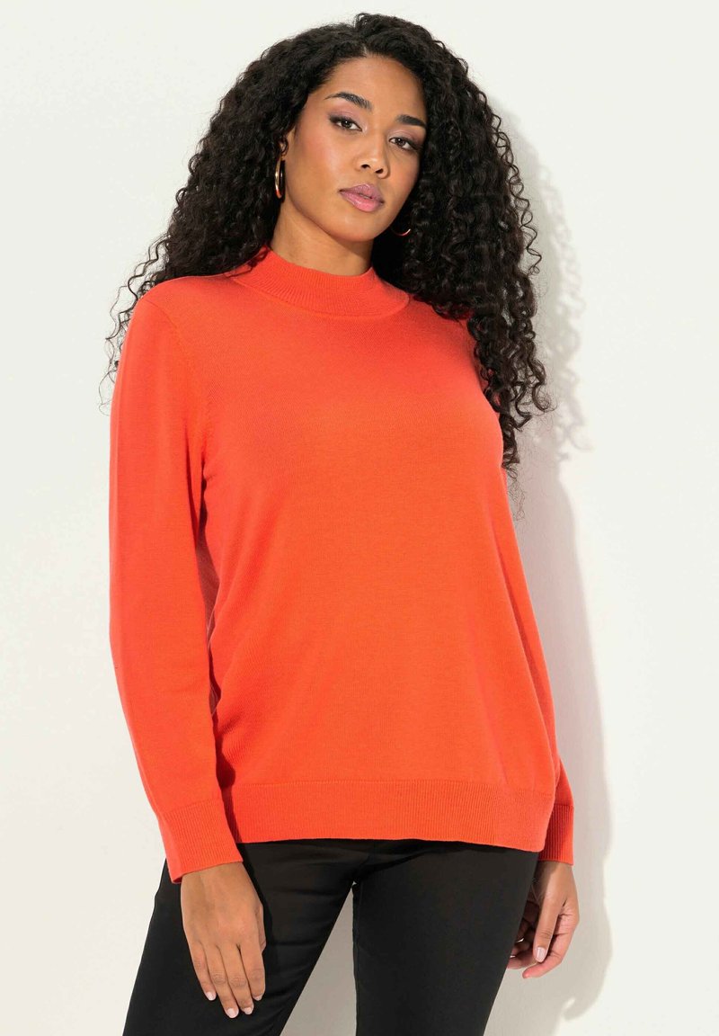 Orange stickad tröja med hög hals, långa ärmar och ribbade muddar; parat med svarta tajta byxor. Slät textur.