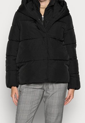 Vrouw met een zwarte pufferjas met capuchon over een beige coltrui, gecombineerd met grijze geruite broek, staand tegen een effen achtergrond.