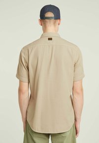 Korte mouwen beige shirt met een achterpaneel, gebogen zoom en labeldetail. Gecombineerd met olijfgroene broek en een blauwe pet. Gladde textuur.