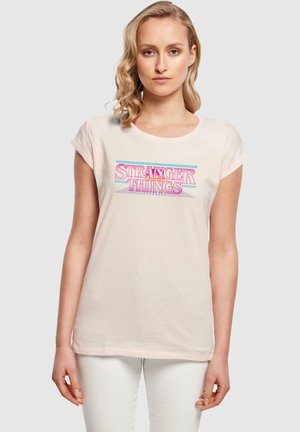 Helles rosa Baumwoll-T-Shirt mit kurzen Ärmeln, das das Logo von "Stranger Things" in buntem, stilisiertem Typografie über die Vorderseite zeigt.