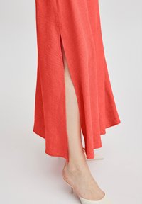 Korallenfarbenes Maxikleid mit seitlichem Schlitz, hergestellt aus leichtem Stoff. Der Saum ist fließend und lässt sich gut mit beigen Absatzmules für einen lässigen Look kombinieren.