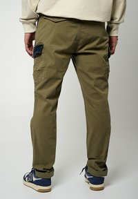 Pantalones cargo color verde oliva con bolsillos laterales y una tela texturizada. La vista trasera muestra piernas ajustadas y acentos de tela contrastantes cerca de los bolsillos.
