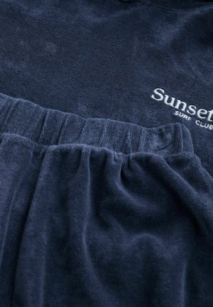 Marinblå frottétyg med en samla midja, prydd med vit broderi som säger "Sunset Surf Club" på den övre sektionen. Mjuk textur.