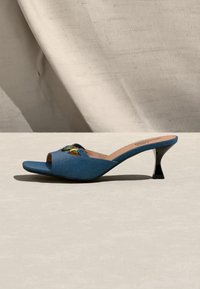 Μπλε mule με ανοιχτό δάχτυλο, διακοσμημένη με χρυσή αγκράφα και καμπυλωτό μαύρο τακούνι kitten, τοποθετημένο σε ανοιχτό μπεζ επιφάνεια μπροστά από υφασμάτινο φόντο.