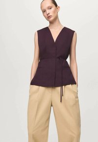 Bezrekawnik o głębokim dekolcie w kształcie litery V, wykonany z ciemnofioletowego materiału, z przewiązanym w talii paskiem, w połączeniu z beżowymi spodniami z wysokim stanem, szerokimi nogawkami i kieszeniami.