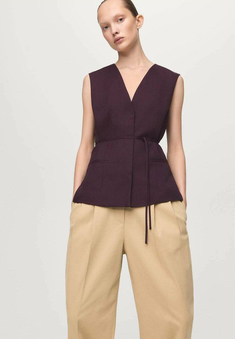 Bezrekawnik o głębokim dekolcie w kształcie litery V, wykonany z ciemnofioletowego materiału, z przewiązanym w talii paskiem, w połączeniu z beżowymi spodniami z wysokim stanem, szerokimi nogawkami i kieszeniami.