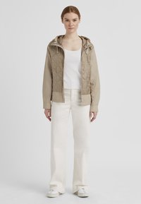 Helle beigefarbene gesteppte Jacke mit Kapuze, ausgestattet mit einem Reißverschluss und zwei seitlichen Taschen, getragen über einem weißen gerippten Oberteil und hellen Jeans.
