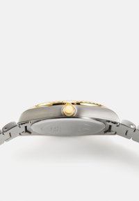 Ceas din oțel inoxidabil cu bezel și coroană accentuate cu aur. Dispune de un capac din argint neted și brățară din linkuri metalice.