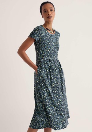 Femme debout les mains dans les poches, portant une robe midi bleue à manches courtes à motif floral et un collier vert, fond neutre.