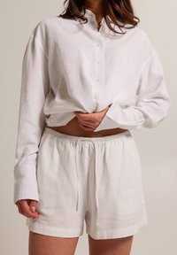 Žena v volné bílé lněné košili na knoflíky a odpovídajících elastických šortkách, drží košili v pase.