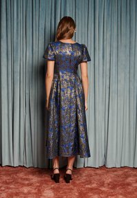 Žena stojaca otočená chrbtom ku kamere, oblečená v modro-zlatej kvetinovej midi šatách a čiernych sandáloch na opätku, na pozadí svetlomodrého závesu.