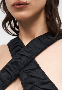 Κοντινό πλάνο ενός ατόμου που φοράει μαύρο ρούχο από ραμμένο ύφασμα που διασταυρώνεται στο στήθος, εμφανίζοντας τα χείλη και μια ελιά πάνω από το χείλος.