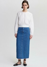 Frau in einer weißen Bluse mit langen Ärmeln, blauer hochtailierter Denim-Midirock mit kleinen Punkten, schwarzen Schnürschuhen, die vor einem schlichten Hintergrund steht.
