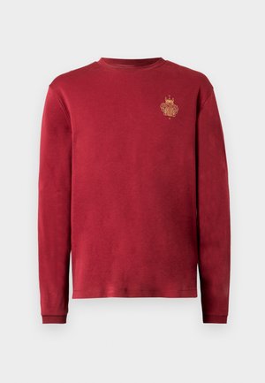 Raudonas ilgomis rankovėmis medvilninis megztinis. Apvalus kaklo iškirptė ir mažas auksinės spalvos logotipo siuvinėjimas kairėje krūtinės pusėje. Lygus paviršius.