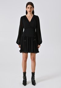 Zwarte jurk met lange, doorschijnende mouwen, een V-halslijn en een aansluitende taille met vetersluiting; gestructureerde stof; gelaagde rok. Zwarte enkelboots.
