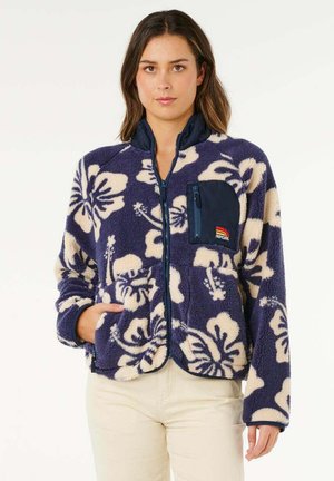 Frau trägt eine marineblaue Fleecejacke mit Reißverschluss, verziert mit cremefarbenen Blumenmustern und einer Brusttasche mit Reißverschluss, kombiniert mit hellfarbenen Hosen.