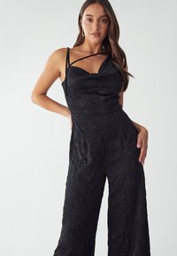 Black jumpsuit with a fitted bodice and wide leg. Features crinkled texture and thin straps, creating a structured design.