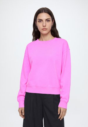 Mujer con cabello largo y oscuro que lleva una sudadera rosa brillante y pantalones negros, mirando hacia adelante con una expresión neutral sobre un fondo liso.