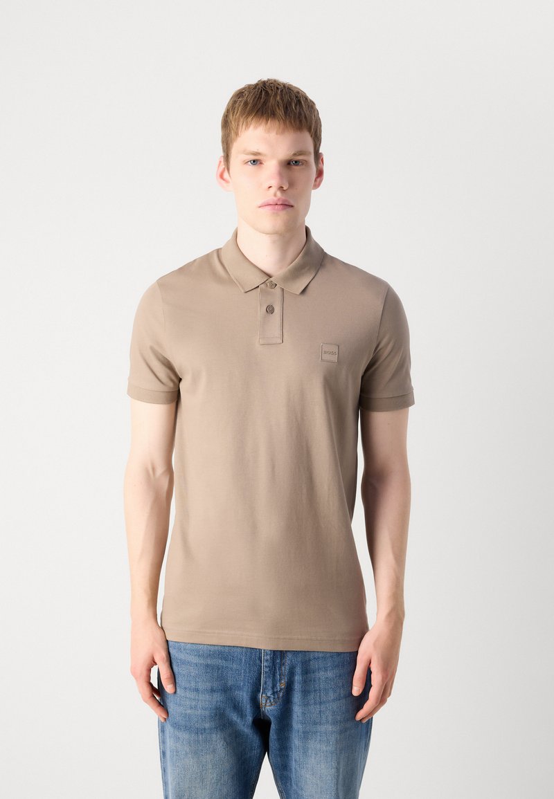 Jeune homme portant un polo beige à manches courtes et un jean bleu, se tenant devant un fond blanc uni.