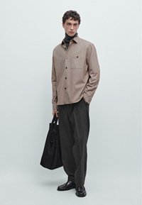 Brun skjorte med knapper og en frontlomme, parret med mørke vide bukser og blanke sorte sko, mens der holdes en sort tote-bag.