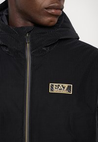 Giacca nera con cappuccio realizzata in tessuto testurizzato, caratterizzata da un'etichetta con logo dorato e una chiusura con zip con zip dorata a contrasto.